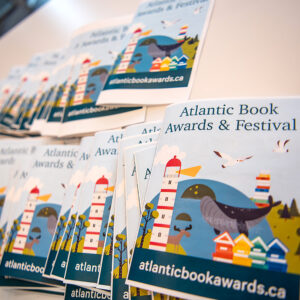 Photo of an array of program booklets for the Atlantic Book Awards Gala from 2025. Happy illustration of lighthouse, water, seagulls and little painted houses on the covers.