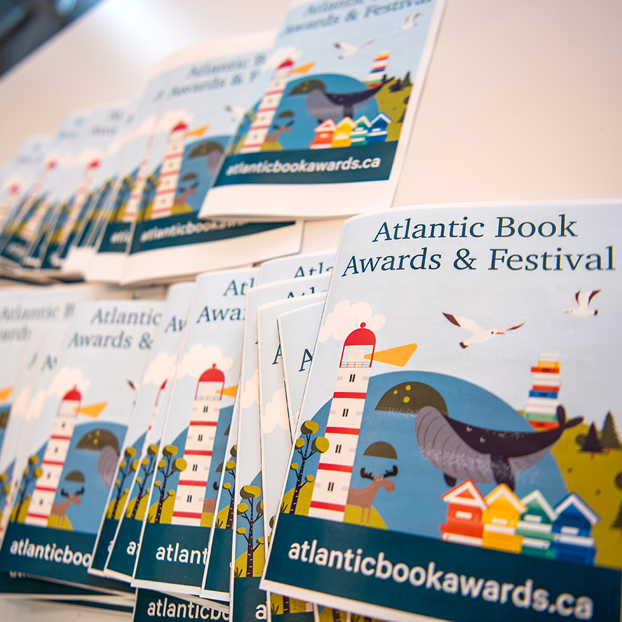 Photo of an array of program booklets for the Atlantic Book Awards Gala from 2025. Happy illustration of lighthouse, water, seagulls and little painted houses on the covers.