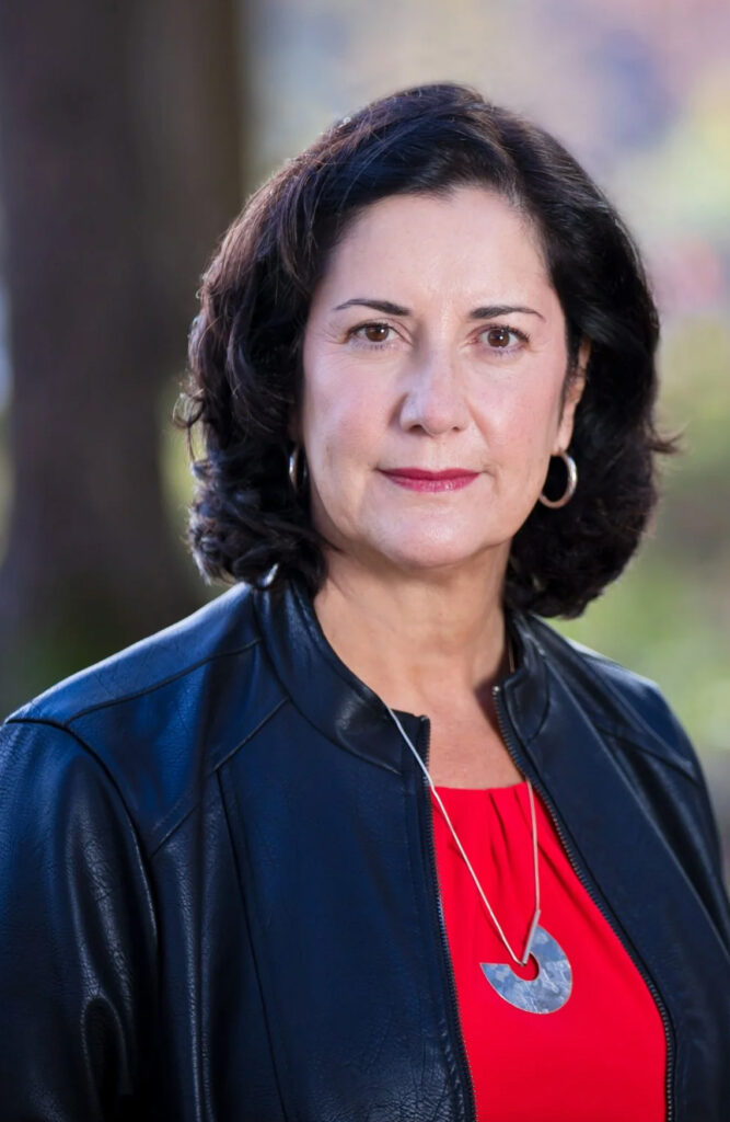 Portrait of dark haired the author with black jacket over red top and a semi-circular disc necklace